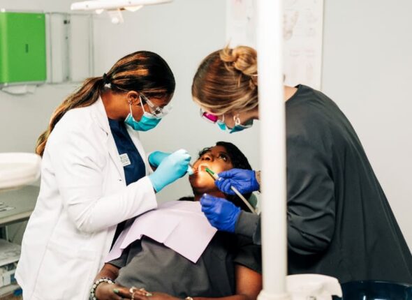 A Dental Assistant Instructor and a Dental Assistant Students Stand Over a Reclined Patient in a Dental Chair, using a suction and tools in the Patient’s Mouth
