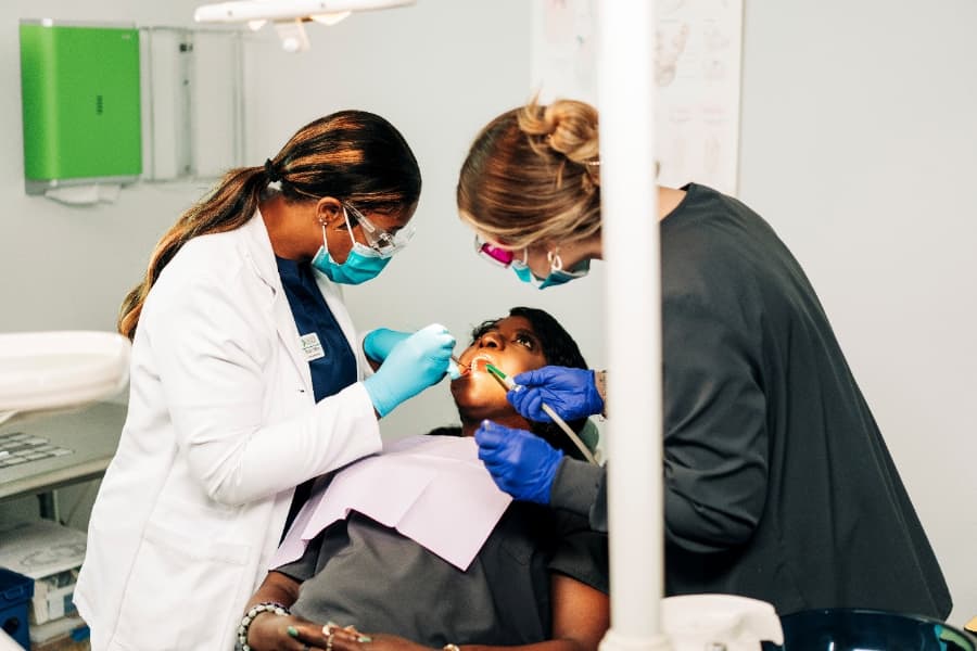 A Dental Assistant Instructor and a Dental Assistant Students Stand Over a Reclined Patient in a Dental Chair, using a suction and tools in the Patient’s Mouth